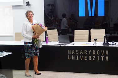 A woman stands holding a bouquet of flowers in a lecture hall at Masaryk University, with rows of seating and a presentation screen in the background. A woman stands holding a bouquet of flowers in a lecture hall at Masaryk University, with rows of seating and a presentation screen in the background.