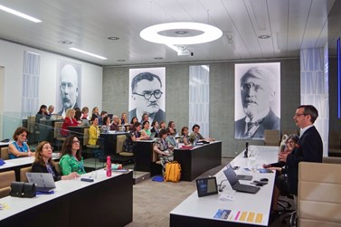 A professional seminar in progress within a modern lecture hall where a presenter is addressing an audience of attentive listeners. The room features stylish decor, including portraits on the walls, contemporary lighting, and equipped with advanced technology for presentations. A professional seminar in progress within a modern lecture hall where a presenter is addressing an audience of attentive listeners. The room features stylish decor, including portraits on the walls, contemporary lighting, and equipped with advanced technology for presentations.