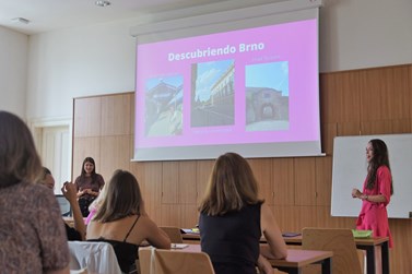Classroom setting with attendees looking towards a presenter in a pink dress giving a presentation about Brno, as depicted on the projector screen showing photos and text. Classroom setting with attendees looking towards a presenter in a pink dress giving a presentation about Brno, as depicted on the projector screen showing photos and text.